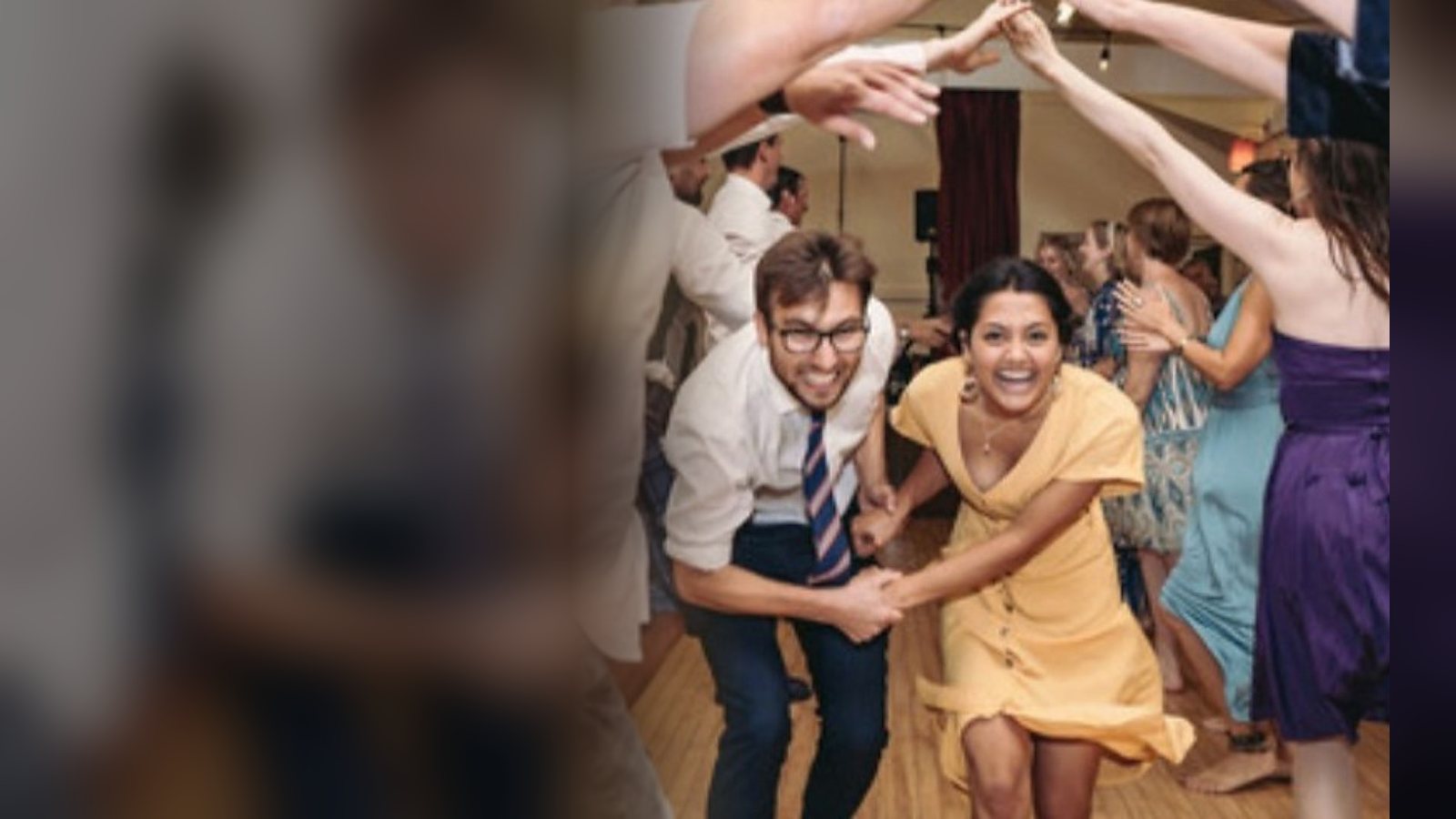 Two wedding guests bursting through an arch of raised hands mid-ceilidh, both laughing, the floor packed behind them