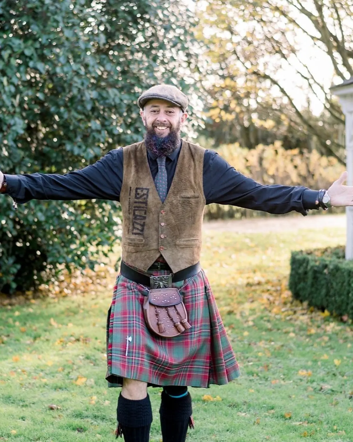 Schuggie in flat cap and kilt, arms thrown wide in welcome in an autumn garden